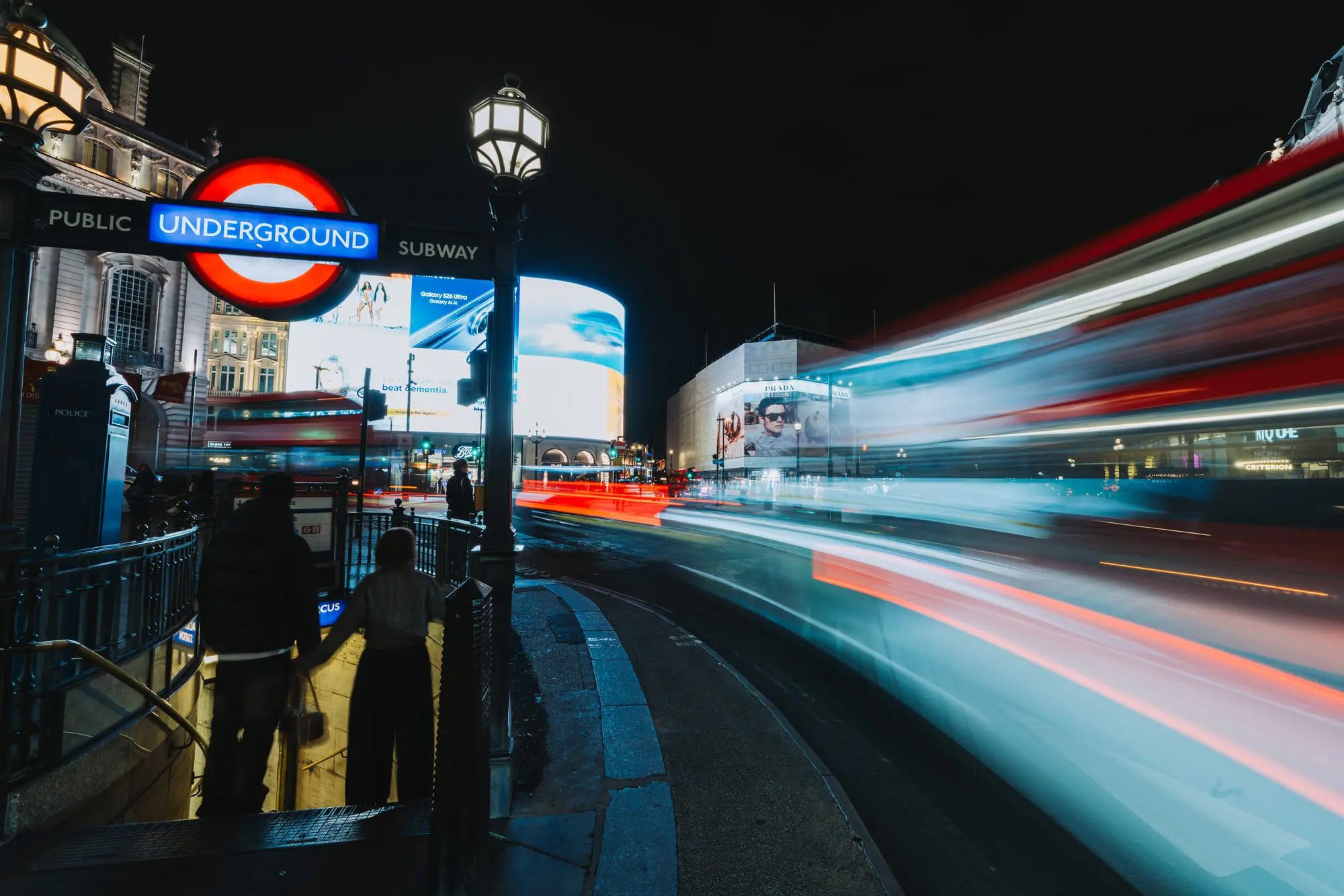Piccadilly Circus at night, the Underground sign glowing as a bus drags light across the frame