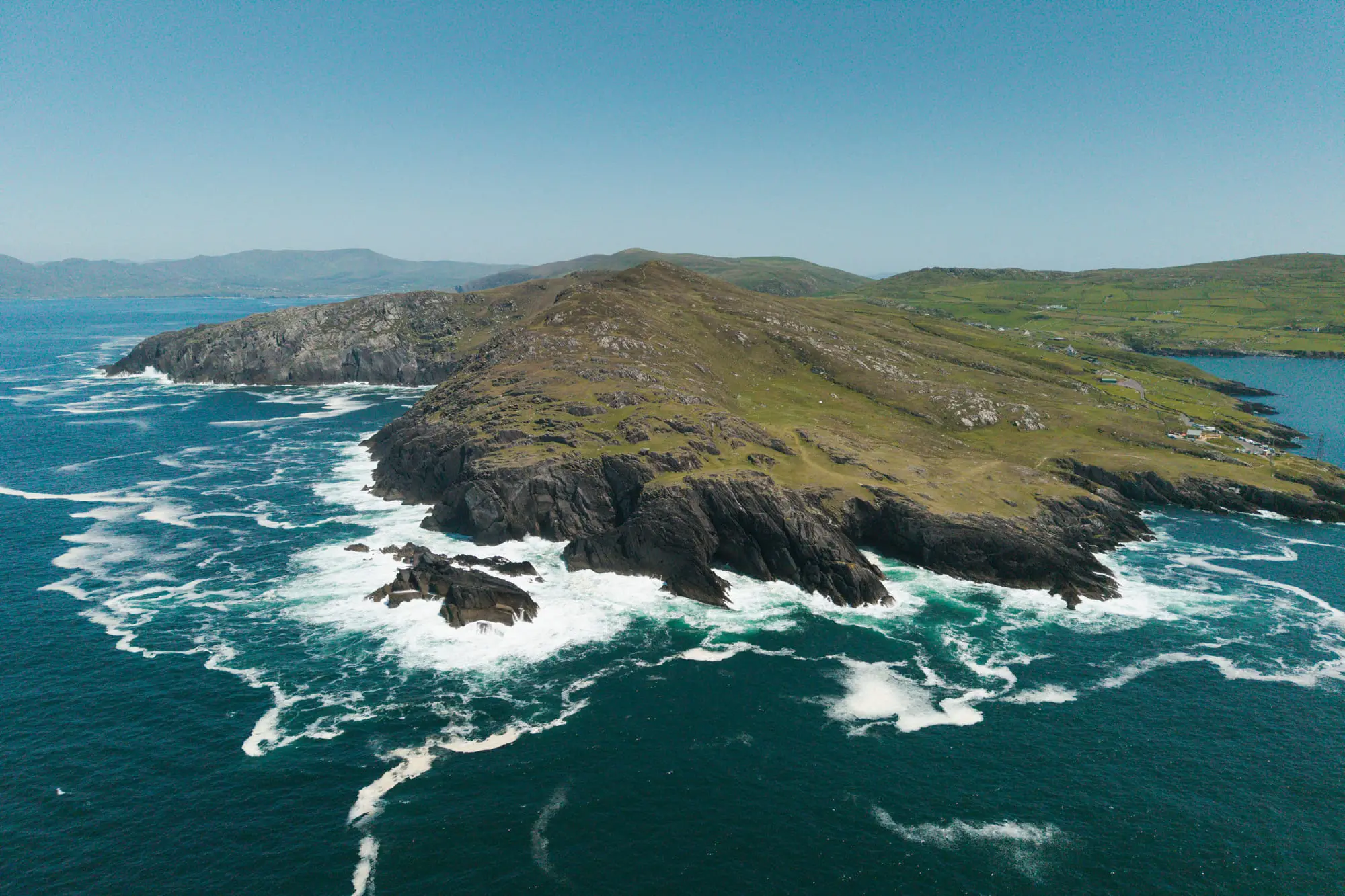 A rugged headland ringed with white surf, from the air