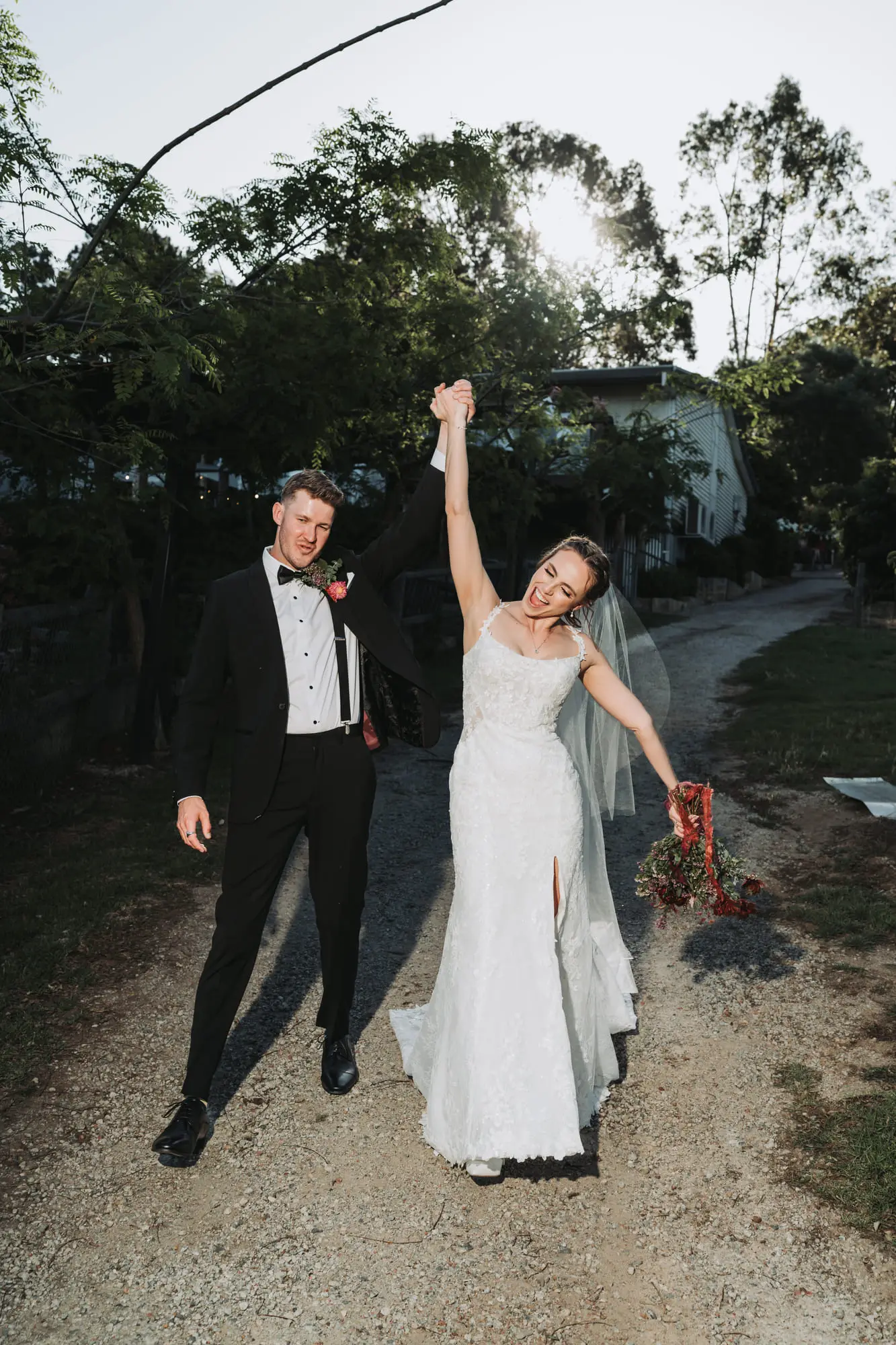 Bride and groom on a path, hands up, bouquet held high, backlit